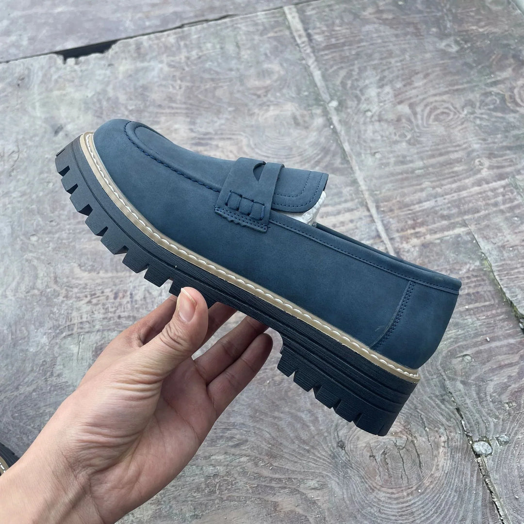Hand holding a navy blue men's casual loafer shoe with chunky black sole on wooden floor