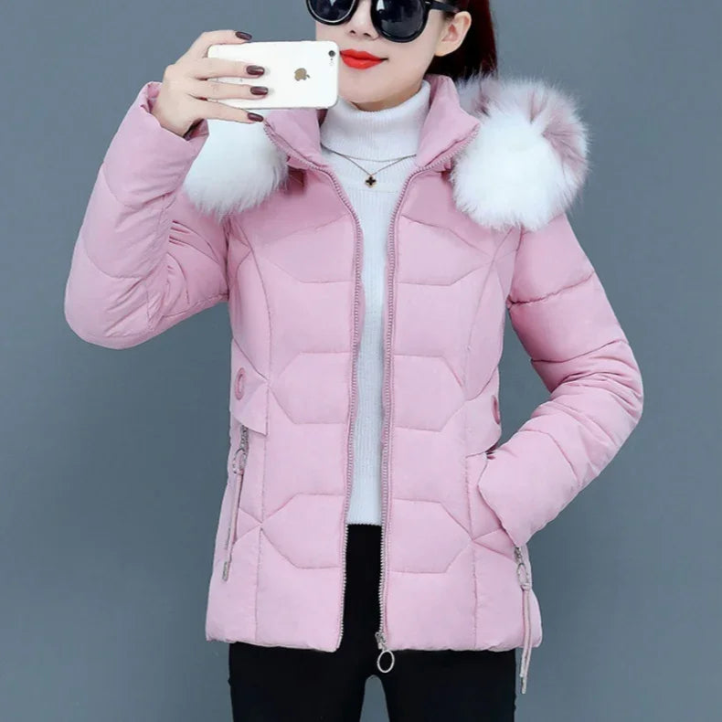 Woman wearing a pink padded winter jacket with white faux fur hood, black pants, and white turtleneck, taking a selfie