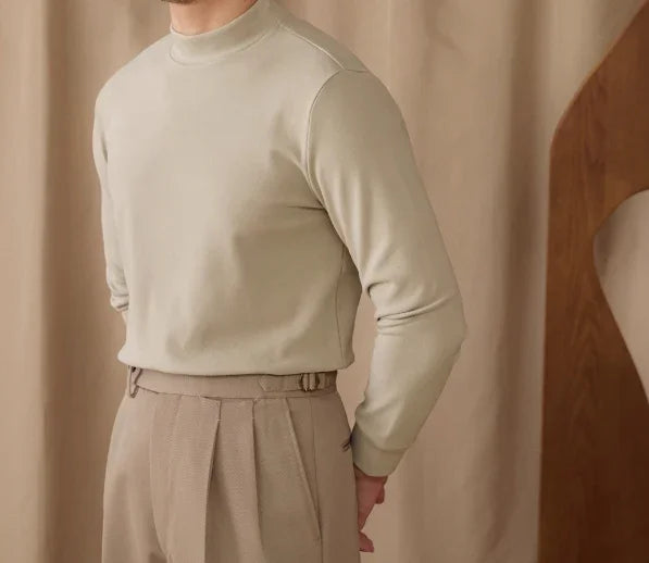 Man wearing beige mock neck sweater and pleated trousers against beige fabric backdrop