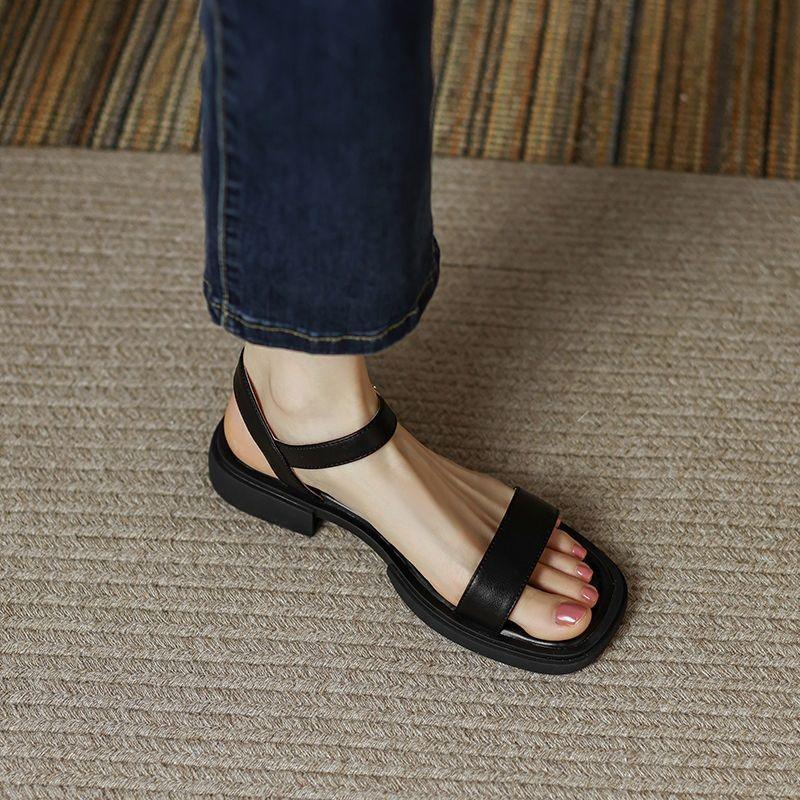 Foot wearing black open-toe leather sandals with ankle strap on textured carpet floor