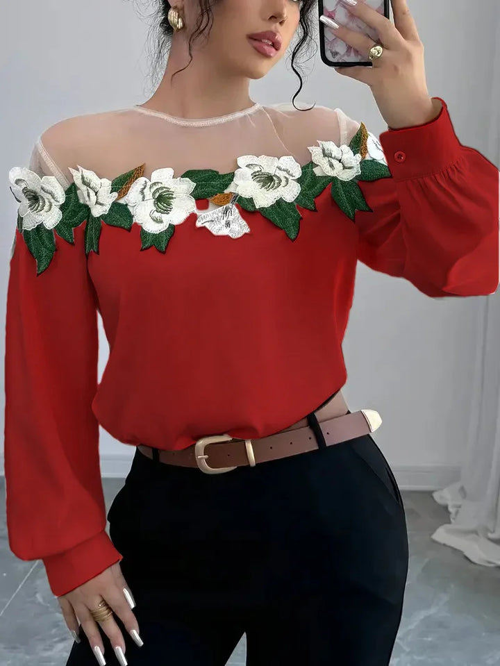 Woman wearing red long-sleeve top with sheer neckline and white floral embroidery, black pants, and brown belt