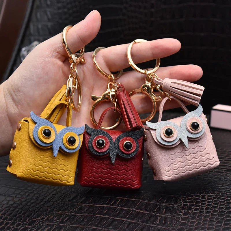 Hand holding three owl-shaped leather keychain bags in yellow, red, and pink with gold rings and tassels