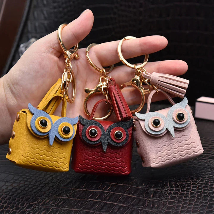 Hand holding three owl-shaped leather keychain bags in yellow, red, and pink with gold rings and tassels
