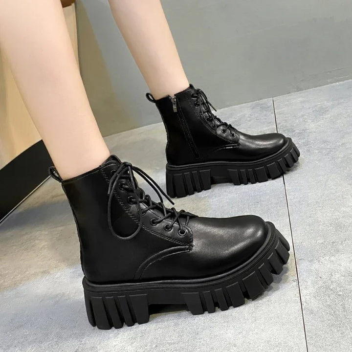 Black leather platform combat boots with chunky lug soles worn on light-toned legs on gray tiled floor