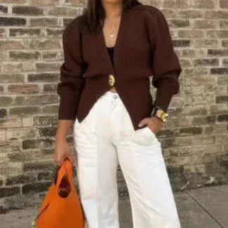 Woman wearing dark brown cardigan with gold buttons and white wide-leg pants, holding orange handbag against brick wall