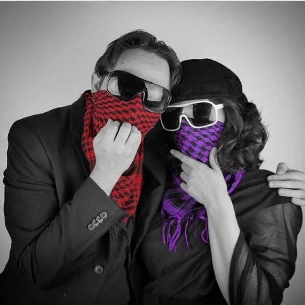 Two people wearing sunglasses and patterned scarves covering their faces, one red and one purple, posed against a plain background