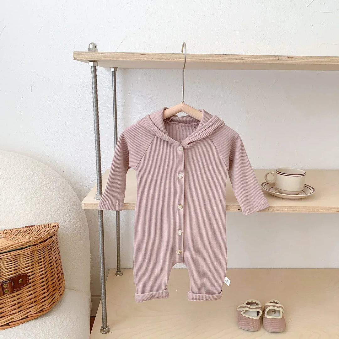 Light pink ribbed baby onesie with hood hanging on wooden shelf near matching baby shoes and wicker basket