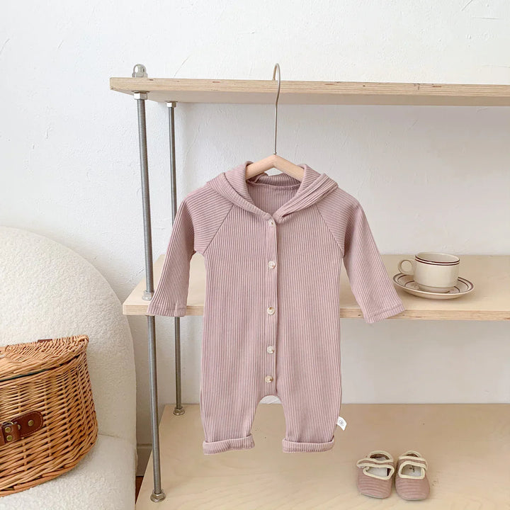 Light pink ribbed baby onesie with hood hanging on wooden shelf near matching baby shoes and wicker basket