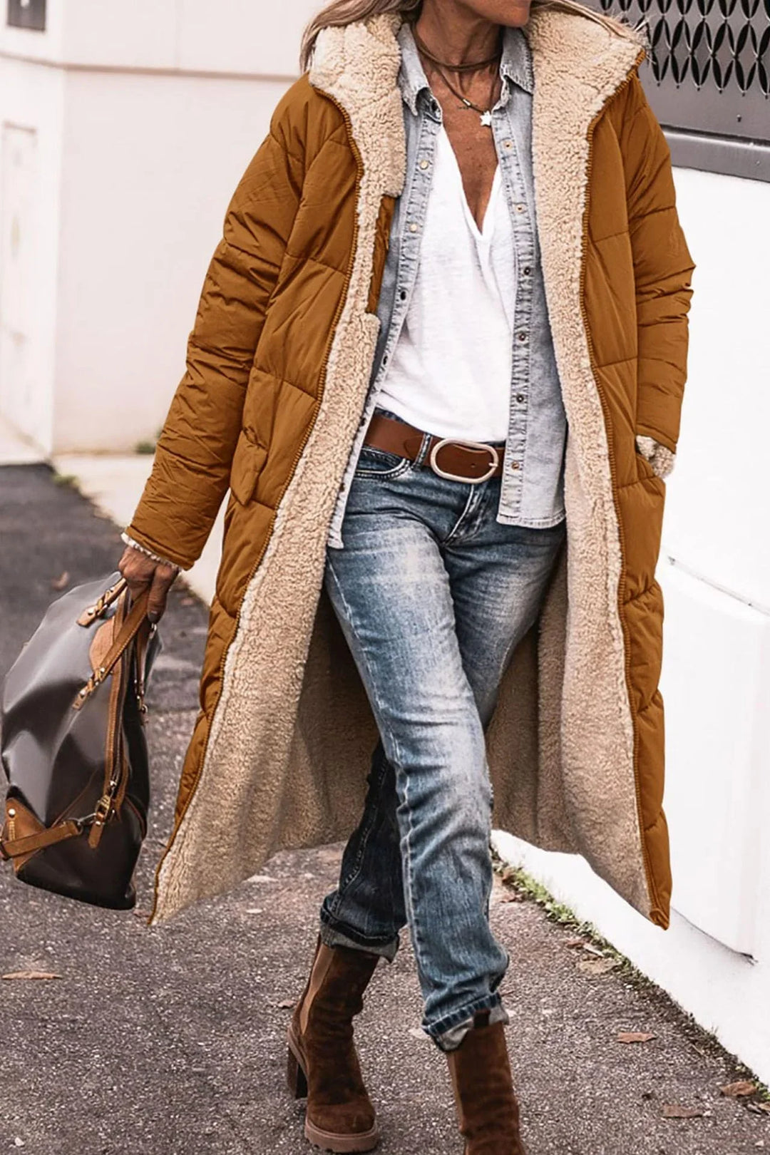 Woman wearing long brown quilted coat with beige fleece lining, white shirt, denim jacket, jeans, and brown boots