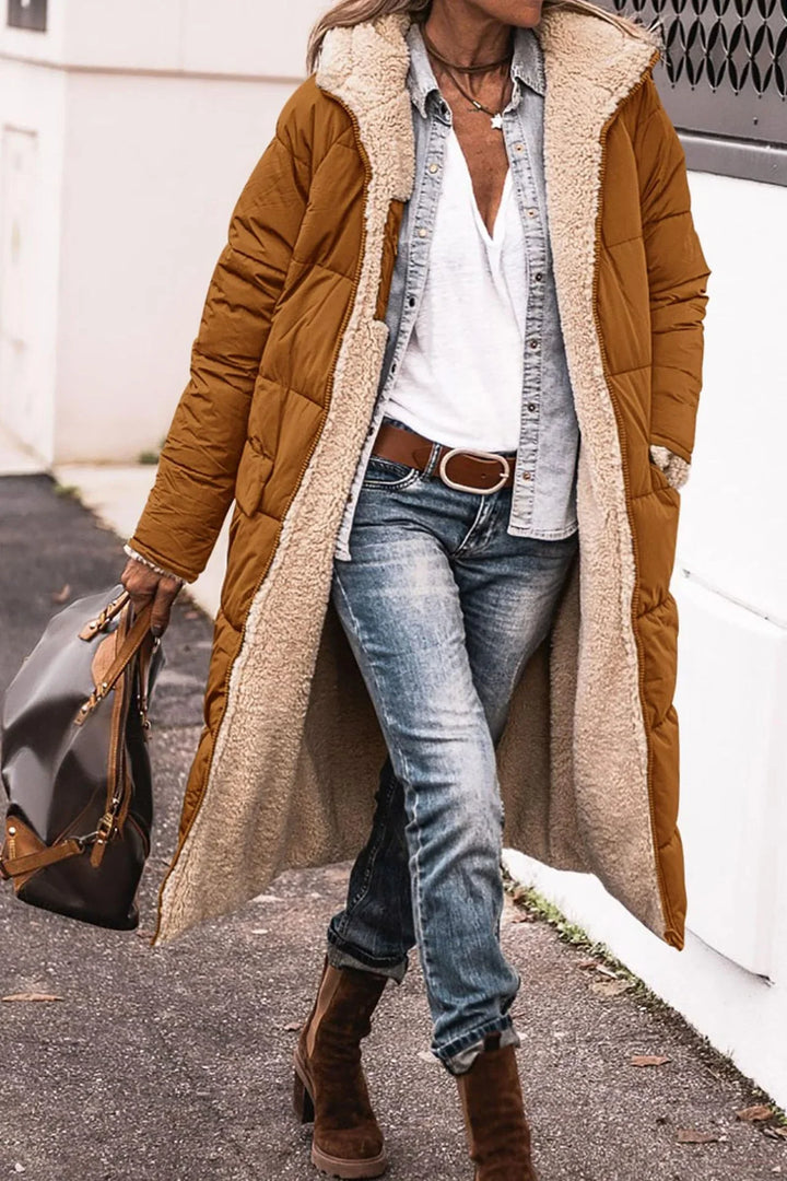 Woman wearing long brown quilted coat with beige fleece lining, white shirt, denim jacket, jeans, and brown boots