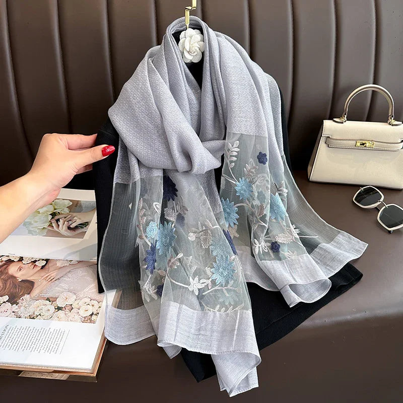 Elegant gray scarf with blue floral embroidery draped on black fabric, styled with beige handbag and sunglasses