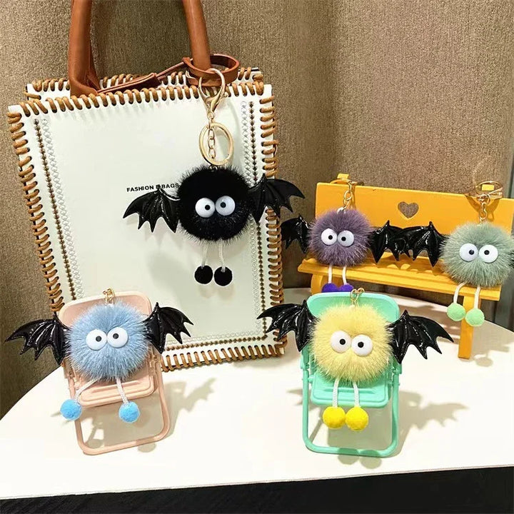 Colorful fluffy bat keychains with big eyes and black wings displayed on mini chairs and a white handbag