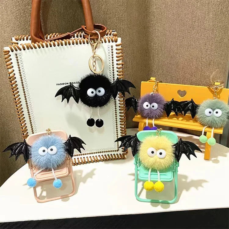 Fluffy keychain charms with big eyes and black bat wings displayed on mini chairs and a white bag with brown handles