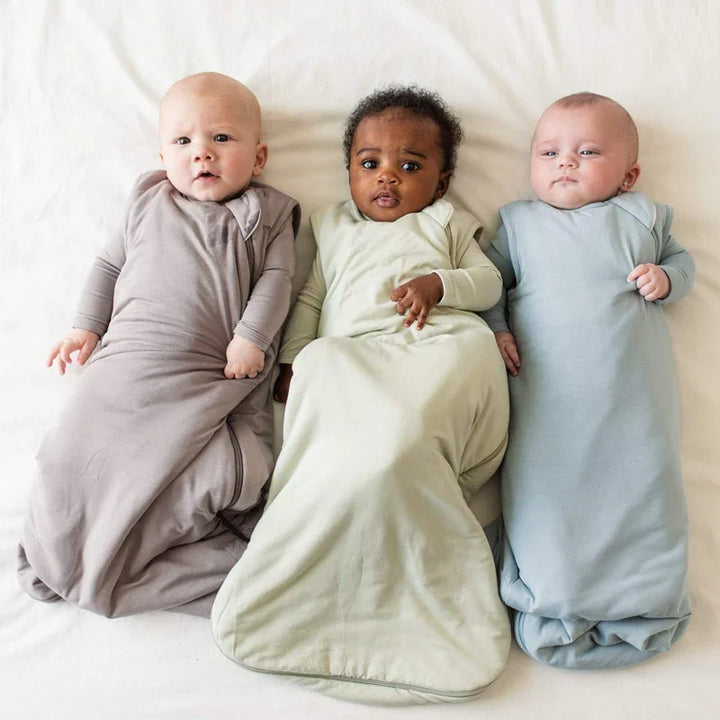 Three diverse babies lying on a white blanket wearing pastel-colored sleep sacks in grey, green, and blue