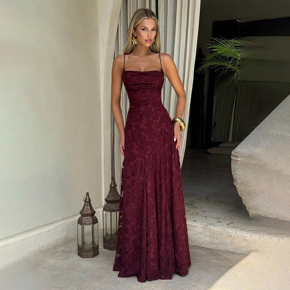 Woman wearing a burgundy floral lace evening gown with spaghetti straps, standing indoors beside decorative lanterns