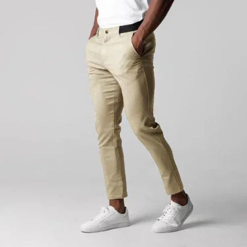 Man wearing beige slim-fit chinos and white sneakers standing against plain gray background