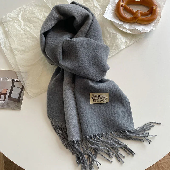 Soft gray fringed scarf with Undreson label on white table next to a soft pretzel and photo card