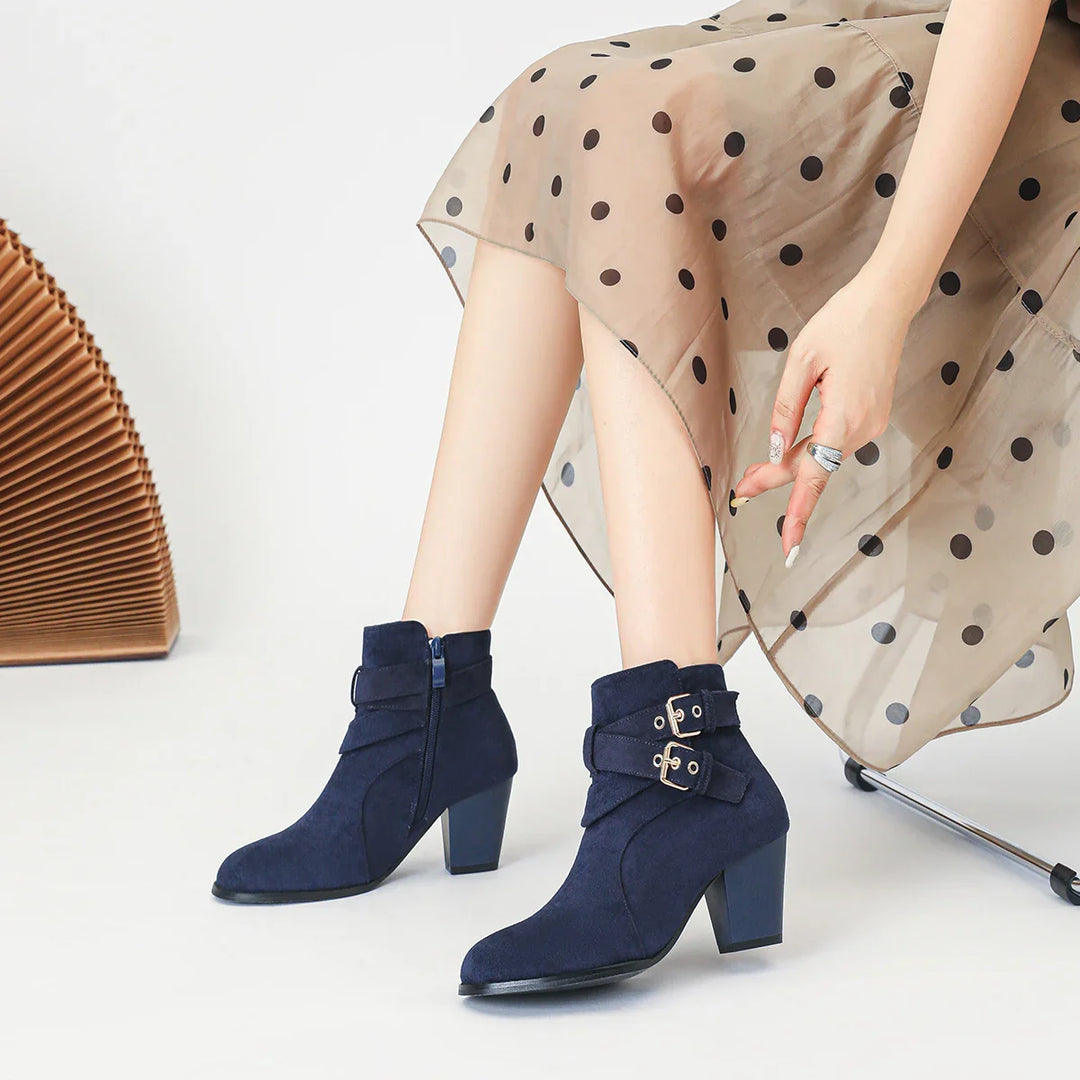 Woman wearing navy suede ankle boots with block heels and double buckle straps, beige polka dot skirt