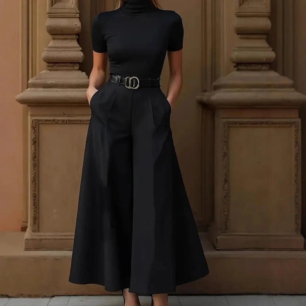 Woman wearing black turtleneck top with matching wide-leg culotte pants and belt standing against ornate beige wall
