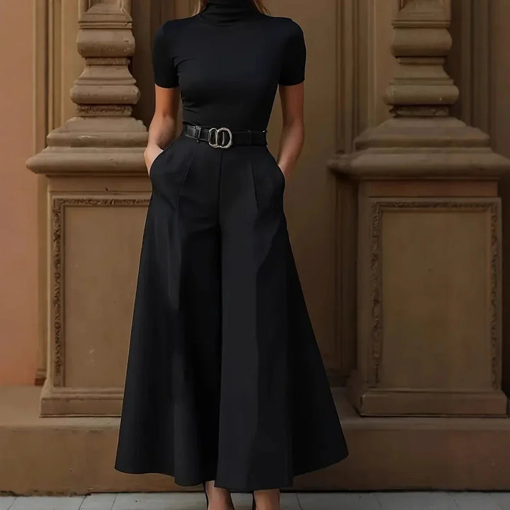 Woman wearing black turtleneck top with matching wide-leg culotte pants and belt standing against ornate beige wall
