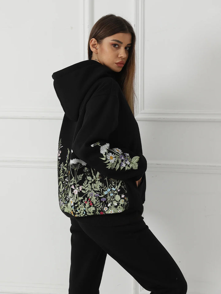 Woman wearing black hoodie with colorful floral embroidery on back and sleeves against white wall