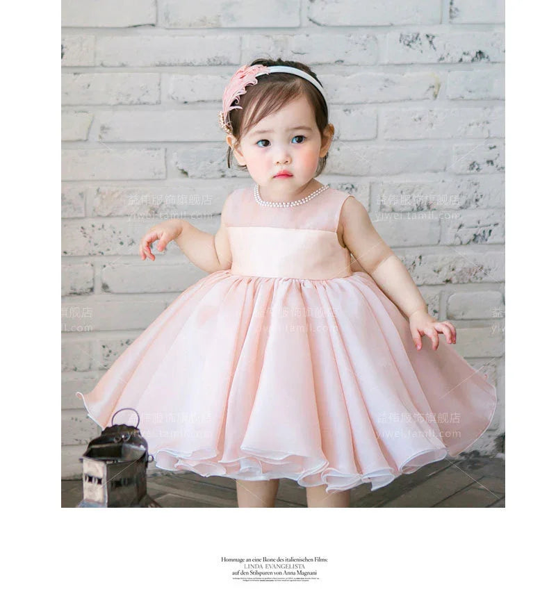 Toddler girl in a light pink sleeveless dress with pearl necklace and floral headband standing indoors