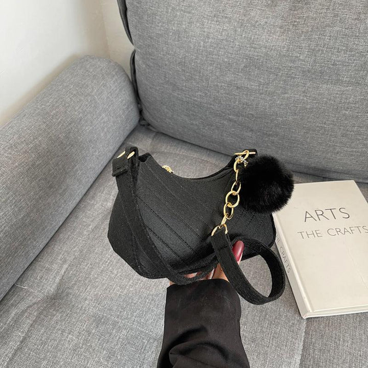 Black quilted handbag with gold chain and black pom-pom, held on grey sofa beside book titled Arts The Crafts