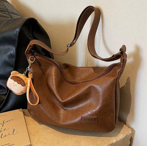Brown leather shoulder bag with adjustable strap and cute plush keychain on beige stone surface