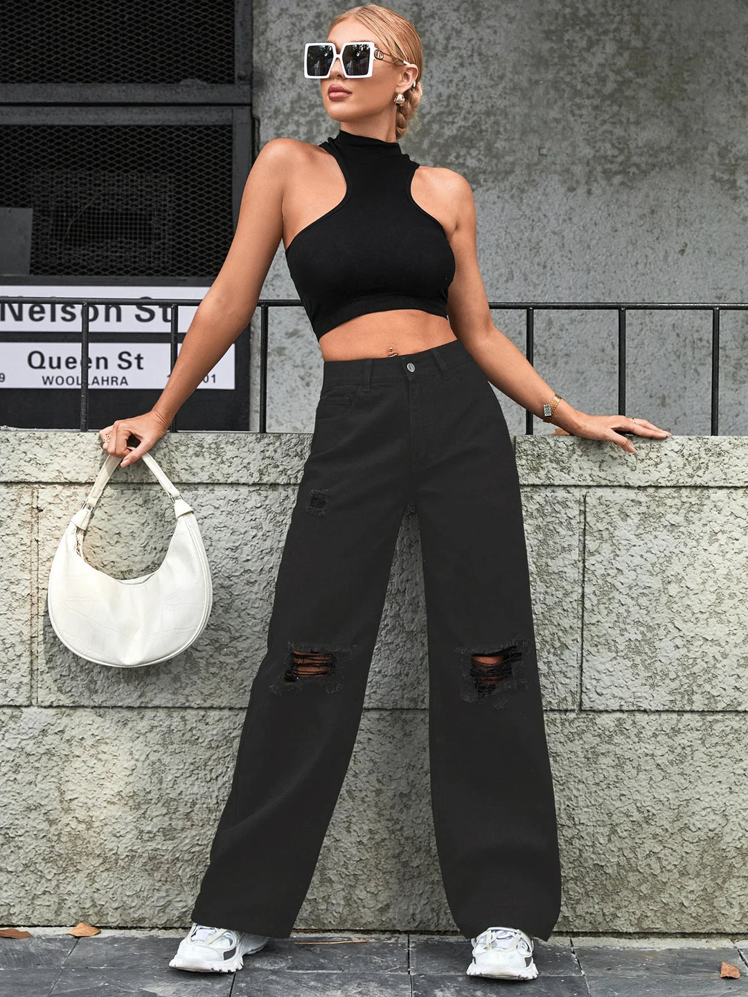 Woman wearing black halter crop top, ripped wide-leg black jeans, white sneakers, holding white handbag outdoors