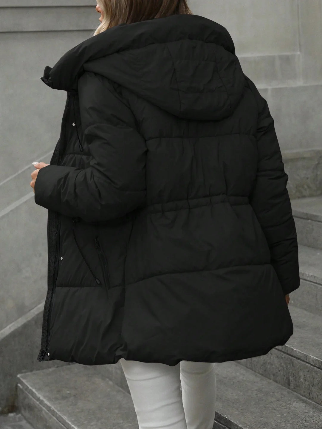 Woman wearing black puffer coat with hood on stone stairs, white pants visible