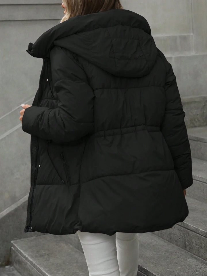 Woman wearing black puffer coat with hood on stone stairs, white pants visible