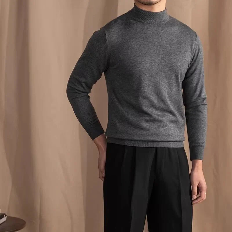 Man wearing charcoal gray mock neck sweater and black pleated pants against beige backdrop