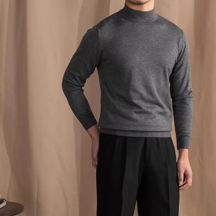 Man wearing charcoal gray mock neck sweater and black pleated pants against beige backdrop