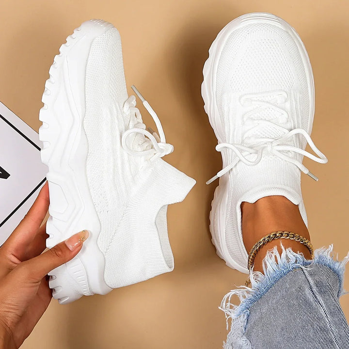 White chunky knit sneakers with thick soles, worn with frayed jeans and gold anklet on beige background