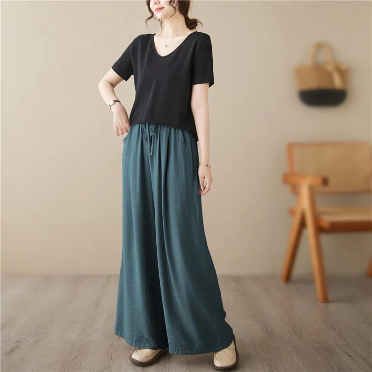 Woman wearing black short sleeve top and teal wide-leg pants in a minimalist indoor setting