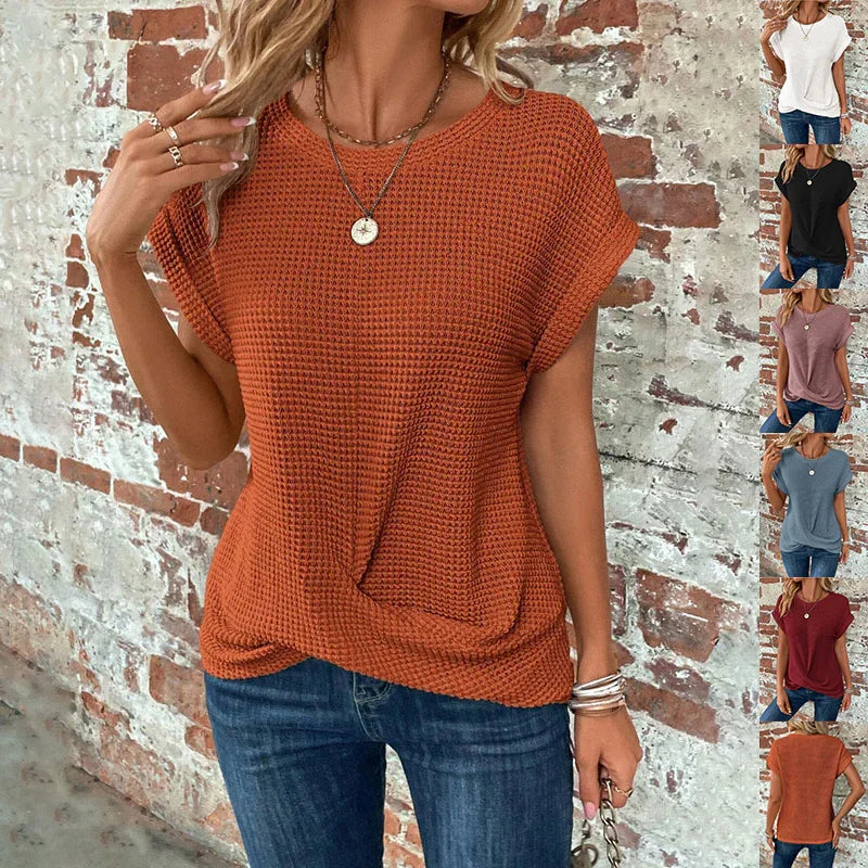Woman wearing rust orange waffle knit short sleeve top with twisted front and blue jeans against brick wall