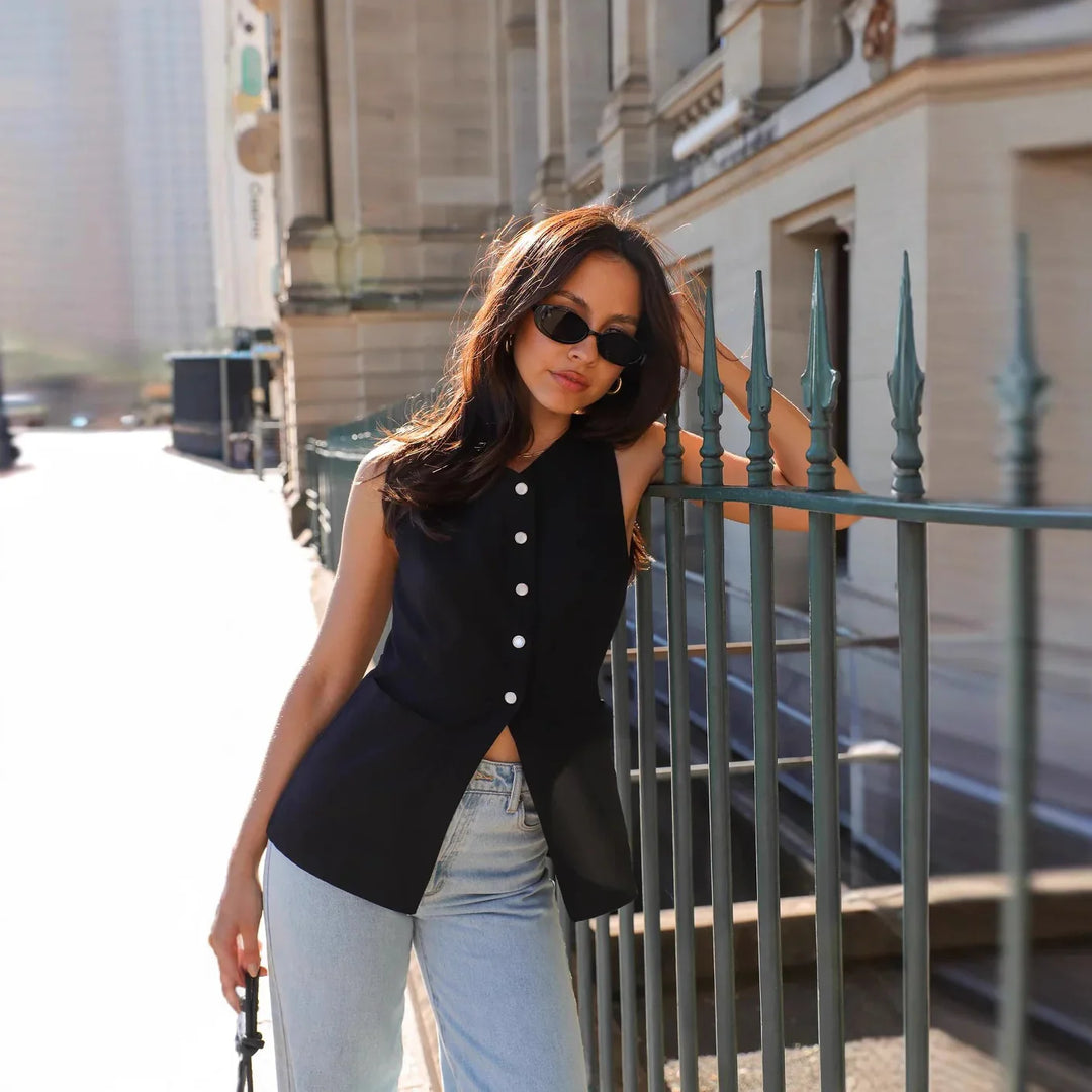 Woman wearing sleeveless black buttoned top and light blue jeans leaning on metal fence in urban setting