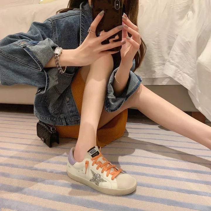 Woman in denim jacket and orange dress sitting on striped carpet wearing white sneakers with star and orange laces