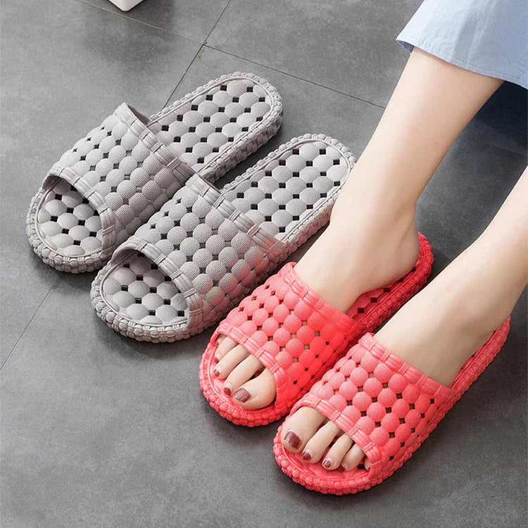 Pair of red open-toe slippers worn on feet beside a pair of gray slippers on gray tile floor