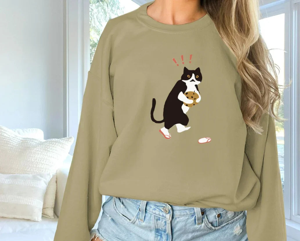 Woman wearing beige sweatshirt with graphic of black and white cat holding cookie indoors