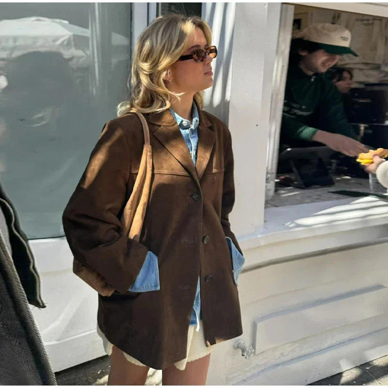 Woman wearing a brown oversized blazer, denim shirt, sunglasses, and a tan shoulder bag standing outdoors
