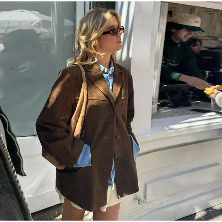 Woman wearing a brown oversized blazer, denim shirt, sunglasses, and a tan shoulder bag standing outdoors
