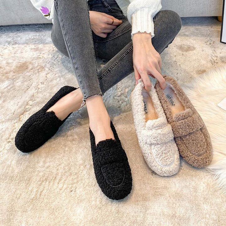 Woman wearing black fuzzy slippers with two pairs of beige and brown fuzzy slippers on carpet
