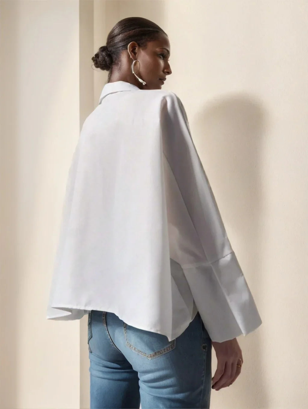 Woman in white oversized blouse and blue jeans standing against beige wall, side profile