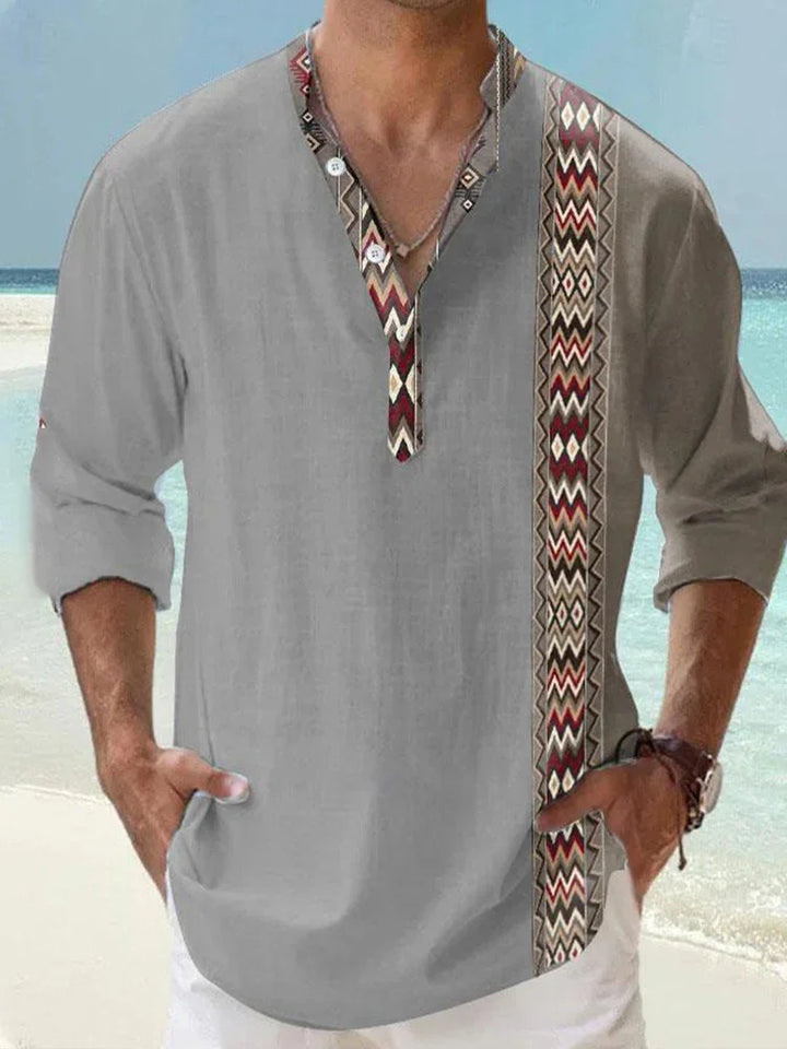 Man wearing light gray long-sleeve shirt with ethnic patterned trim, standing on beach near ocean
