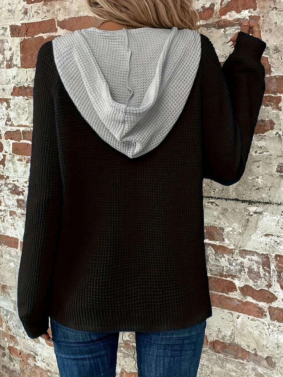 Back view of woman wearing black waffle knit hoodie with gray hood against rustic brick wall