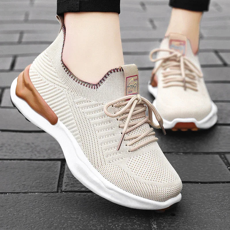 Light beige knit sneakers with white soles and brown accents worn on black pants on gray brick pavement