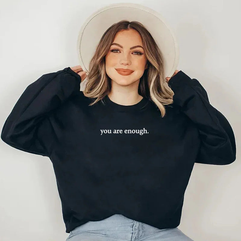 Smiling woman wearing a black sweatshirt with 'you are enough.' text and a beige hat