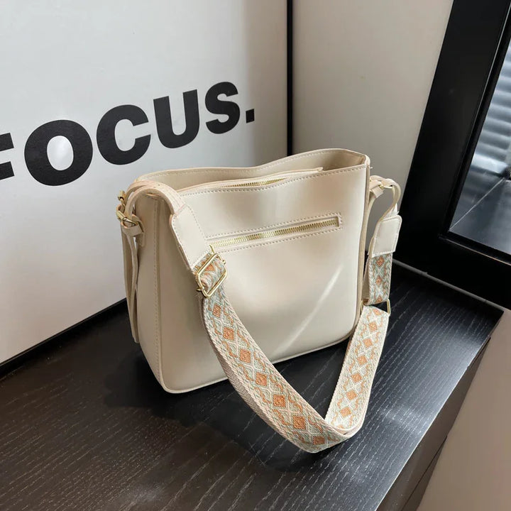 Cream leather handbag with patterned adjustable strap on black wooden surface near window and 'FOCUS' wall art