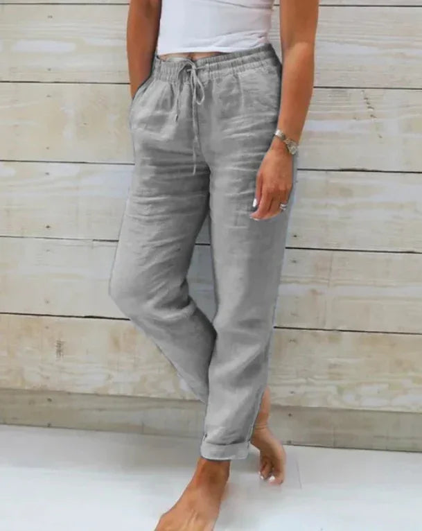 Woman wearing light gray drawstring linen pants with rolled cuffs, standing barefoot against a wooden wall